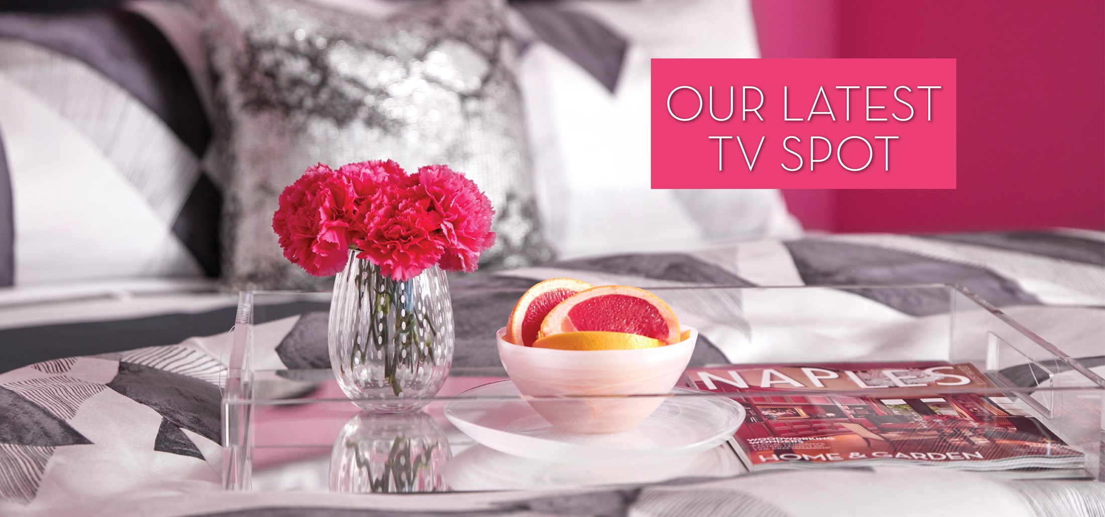 Photo of a tray with flowers and fruit on a bed with black and white bedding. Text: Our Latest TV Spot. Links to TV Spot on Youtube.
