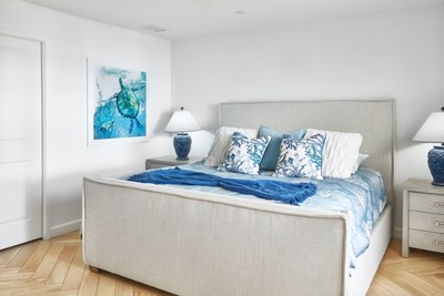 Image of a light grey upholstered bed with blue and white bedding, and sea turtle art on the wall.