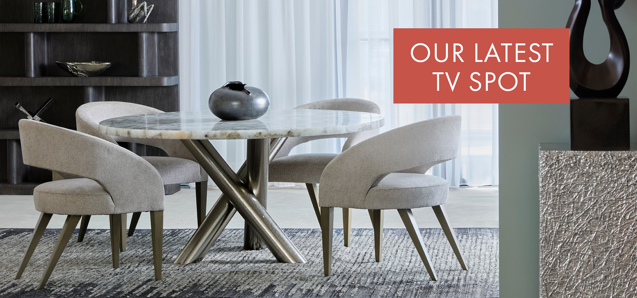 Image of a modern dining table with metal base and upholstered chairs. Text on orange rectangle: Our Latest TV Spot. Links to TV Spot on Youtube.